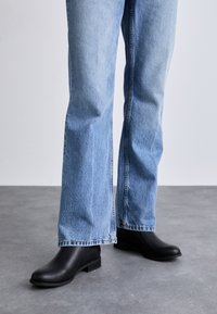 Bottines en cuir noir avec une finition texturée, talon bas en bloc, associées à un jean en denim bleu clair à jambes larges.