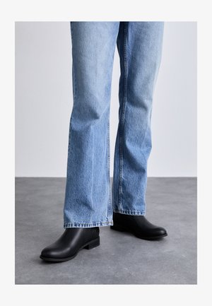 Botas de tornozelo em couro preto com um acabamento texturizado, salto bloco baixo, combinadas com jeans de ganga de perna larga em azul claro.