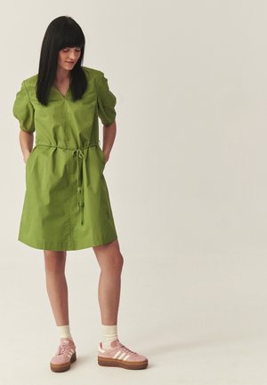 Kobieta o czarnych włosach, ubrana w zieloną sukienkę do kolan z bufiastymi rękawami, przewiązaną w talii, białe skarpetki i różowe trampki Adidas.