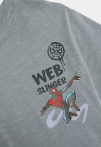 Camiseta gris de manga corta con cuello redondo. Muestra un gráfico colorido de Spider-Man, el texto dice "WEB SLINGER". Tejido suave y texturizado.