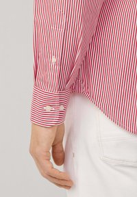 Poignet d'une chemise à manches longues rayée rouge et blanche avec des boutons sur le bras d'une personne, à côté de la poche d'un pantalon blanc et de sa main.
