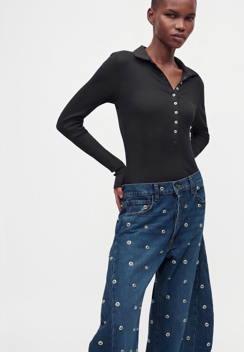 Personne portant un haut noir ajusté à manches longues boutonné et un jean bleu ample avec des clous métalliques circulaires sur un fond clair uni.