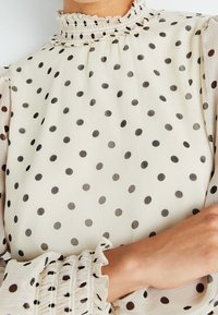 Blusa crema con pois neri; presenta un collo alto con volant e polsini aderenti con bordo a volant, realizzata in tessuto leggero.
