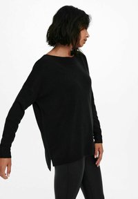 Pull en maille noire avec un col large, des manches longues, des fentes latérales et une coupe décontractée. Matériau texturé et design minimaliste.