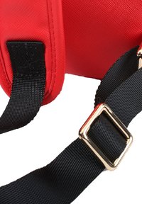 Sac en tissu rouge avec une sangle en nylon noir et des éléments en or. Surface texturée avec un dessus rembourré et un design linéaire pour le confort.