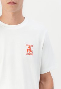 White cotton t-shirt featuring an orange graphic print that reads "SHANGHAI CHAMPION" alongside a boxing figure on the left chest.