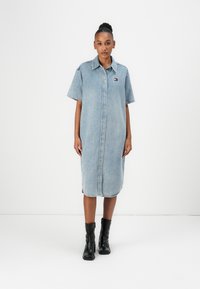 Denim skjortklänning i ljusblå, knälång med korta ärmar, framknäppt och med en liten logotyp på bröstet. Bärs med svarta ankelskor.