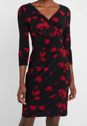 Czarna sukienka do kolan z czerwonym kwiatowym wzorem, z rękawami o długości do łokcia i dekoltem w kształcie litery V, noszona przez osobę z splecionymi rękami.