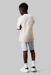 Camisa de algodón con bloques de color crema y beige, combinada con pantalones cortos de mezclilla claros y zapatillas blancas con acentos negros.