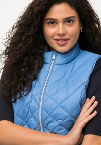Gilet trapuntato blu chiaro con colletto alto e zip frontale; presenta una texture liscia e un motivo a rombi; indossato sopra una camicia scura.