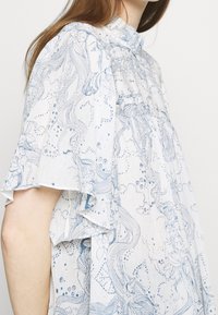 Blusa bianca fluida con motivi floreali blu e linee artistiche, maniche corte con bordi arricciati e dettaglio di scollatura arricciata.