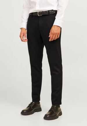 Uomo che indossa pantaloni slim fit neri, cintura in pelle nera, scarpe nere stringate e camicia bianca a maniche lunghe, in piedi davanti a uno sfondo semplice.