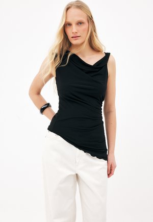 Femme blonde portant un haut noir asymétrique froncé sans manches et un pantalon blanc, posant avec la main sur la hanche devant un fond uni.