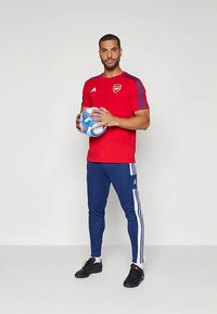 Červené športové tričko s modrými prvkami, v kombinácii s modrými teplákmi s bielymi pruhmi; drží futbalovú loptu s vzorom; čierne topánky.