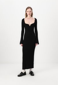 Abito nero in maglia con scollatura profonda, maniche lunghe e polsini a campana. Presenta un accento decorativo dorato sul petto.