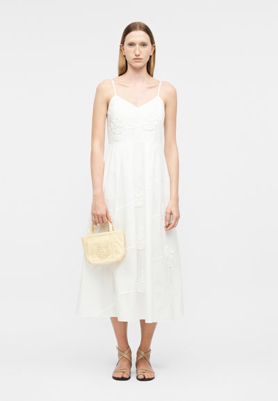 Femme en robe midi blanche sans manches avec broderie florale, tenant un petit sac à main beige et portant des sandales à brides.
