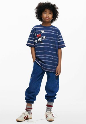 Mėlynos juostelėmis didelio dydžio marškinėliai su animacinio stiliaus grafika ant krūtinės, derinami su laisvo silueto džinsiniais kelnėmis su kišenėmis ir elastingais rankogaliais. Balti sportbačiai su raudonomis detalėmis.