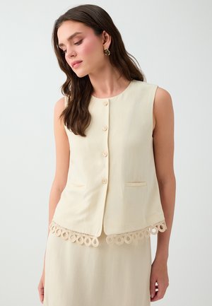 Femme aux longs cheveux foncés porte un haut crème sans manches boutonné avec une bordure en dentelle bouclée décorative et une jupe assortie sur fond uni.