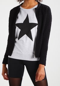 Sort quiltet jakke med lynlås foran, båret over en grå sweatshirt med en stor sort stjerne. Sorte sportsshorts er synlige nedenfor.