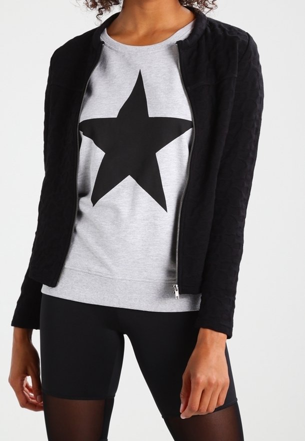 Sort quiltet jakke med lynlås foran, båret over en grå sweatshirt med en stor sort stjerne. Sorte sportsshorts er synlige nedenfor.
