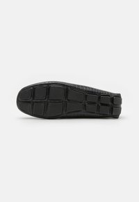 Chaussure plate noire avec une tige en cuir crocodile synthétique texturé. La semelle présente un design de grip à motifs pour traction et durabilité.