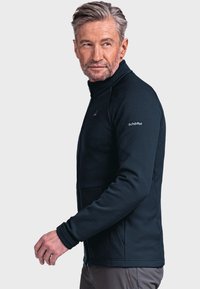 Schwarze Fleecejacke mit hohem Kragen, ausgestattet mit einem Reißverschluss und zwei Fronttaschen. Gerippte Textur und Logo auf der Schulter.