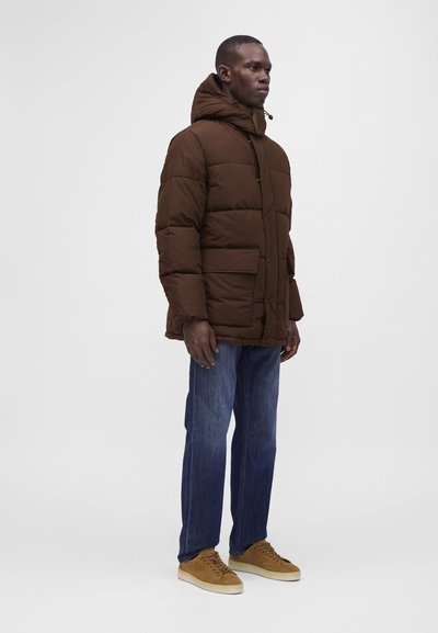 Homme portant une doudoune à capuche marron, un jean bleu et des baskets beige, debout de profil devant un fond uni.