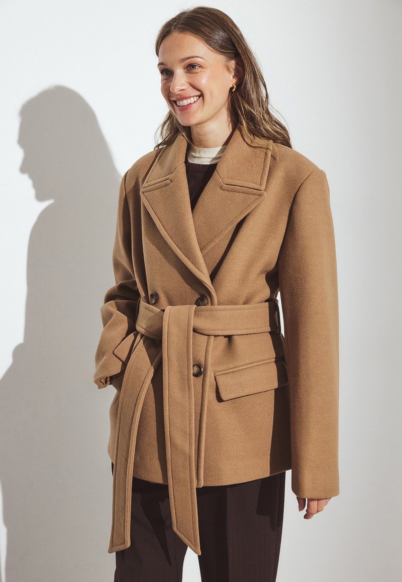 Manteau ceinturé en mélange de laine beige avec un design à double boutonnage, grands revers, poches avant, et une texture lisse, porté sur un haut sombre.
