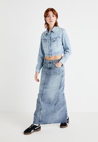 Giacca di jeans azzurro chiaro con design corto e due tasche sul petto, abbinata a una gonna di jeans azzurro chiaro a vita alta e a lunghezza intera. Sneakers nere.