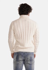 Maglione a maglia color crema con colletto alto, caratterizzato da righe verticali e dettagli a trecce sulle maniche. Indossato con jeans blu sbiaditi.