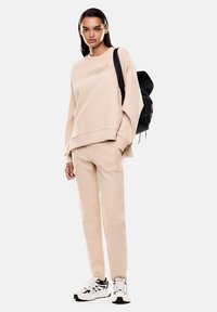 Completo di felpa e pantaloni athleisure beige, in materiale misto cotone, vestibilità rilassata, dettaglio logo testurizzato, indossato con uno zaino nero e sneakers bianche.