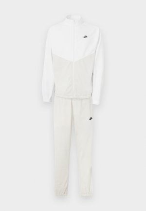 Vit Nike-träningsdräkt med en dragkedjejacka med chevrondesign och byxor med elastisk midja. Inkluderar svarta logotypdetaljer.