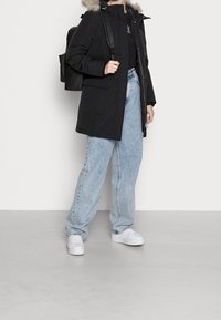 Svart vadderad jacka med fuskpäls på huvan, tillsammans med ljusblå baggy jeans och vita sneakers, samt en svart ryggsäck.