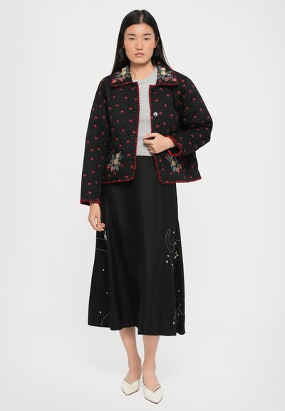 Jachetă neagră matlasată cu cusături roșii și broderie florală, purtată peste un top gri, asortată cu o fustă lungă neagră cu modele celeste.