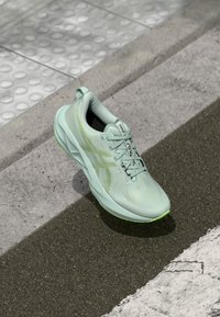 Scarpa da corsa verde chiaro con tomaia testurizzata, lacci e dettagli riflettenti. Presenta un intersuola ammortizzata e una suola in gomma per una maggiore trazione.