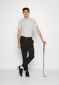 Grå polo skjorte med logo, svarte bukser og svarte joggesko. Figuren holder en golfklubbe og poserer mot en ensfarget bakgrunn.
