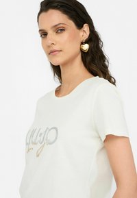T-shirt blanc avec un col rond, à manches courtes, présentant un motif pailleté argenté et doré. Le mannequin porte de grandes boucles d'oreilles en forme de cœur doré.