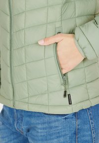 Ljusgrön, vadderad jacka med dragkedjefickor och mjukt material. Har en tagg med "comfortemp" nära fållen.