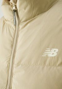 Veste matelassée beige avec fermeture éclair devant et logo New Balance blanc sur la poitrine.