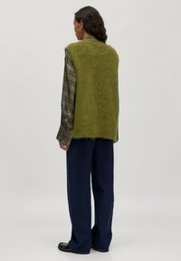 Groene, fuzzy vest over een geruit shirt, gedragen met donkerblauwe wijde broekspijpen. De zachte textuur en gelaagde stijl creëren een contrasterende look.