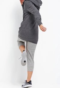 Persona vestida con sudadera gris, pantalones cortos gris claro y zapatillas negras, levantando una pierna mientras se equilibra contra un fondo blanco.