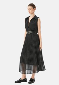 Schwarzes ärmelloses Kleid mit Kragen, Vorderknöpfen, plissiertem Rock und Gürtel. Es hat ein gepunktetes Muster und eine fließende Textur.