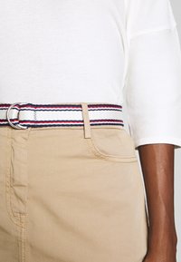 Top de mezcla de algodón en color crema y pantalones beige, que cuentan con un cinturón de tela a rayas con líneas rojas, blancas y azules y una hebilla circular plateada.