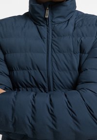 Tummansininen toppatakki korkealla kauluksella, jossa on pitsikuvioita ja vetoketjusuljenta. Materiaali näyttää olevan teksturoitua ja eristettyä.