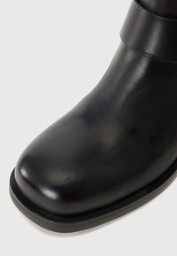 Bota de tobillo de cuero negro con punta redondeada y textura suave. Cuenta con una suela resistente y una banda ancha en la parte superior para un ajuste seguro.