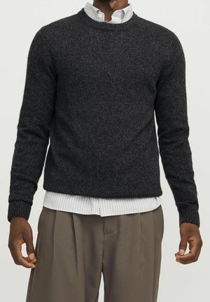 Mand iført mørkegrå strikket sweater over hvidt, stribet skjorte og løse brune bukser, stående foran en ensfarvet baggrund.