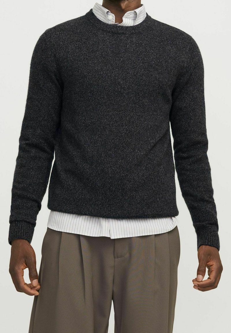 Homme portant un pull en maille gris foncé sur une chemise blanche rayée et un pantalon marron ample, debout devant un fond uni.