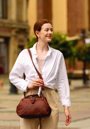 Brown leather handbag with a rounded shape, featuring a buckle closure and a detachable strap, paired with a white shirt and beige pants.