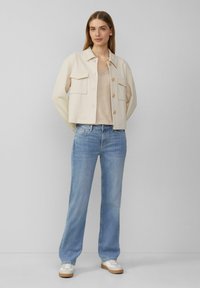 Vrouw die met de handen op de rug staat, gekleed in een beige colbert, gestreepte beige top, lichte blauwe jeans en witte sneakers tegen een effen achtergrond.