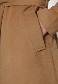 Kamelfarbenes Wollcape mit weicher Textur, breitem Kragen und Seitentaschen sowie einem gürtelten Design für eine verstellbare Passform.
